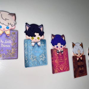 May include: A collection of five decorative bookmarks featuring cartoon cat characters perched atop miniature book replicas. The books have titles like "The Little Prince" and "Dracula", with various colors and designs.