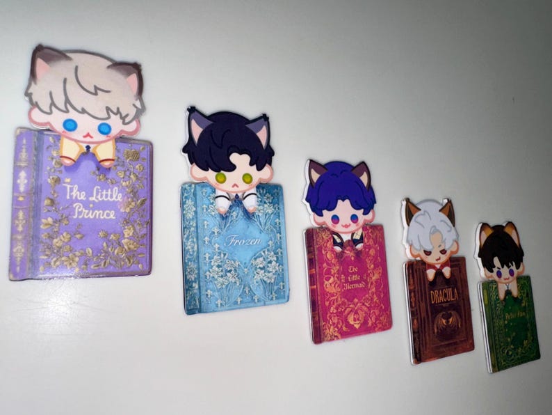 May include: A collection of five decorative bookmarks featuring cartoon cat characters perched atop miniature book replicas. The books have titles like "The Little Prince" and "Dracula", with various colors and designs.