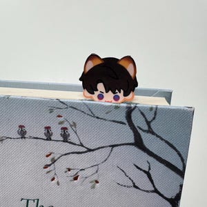 May include: A black and brown anime-style bookmark in the shape of a character's head, placed on top of a book. The book has a light blue cover with a tree branch design and the title "The Travelling Cat".