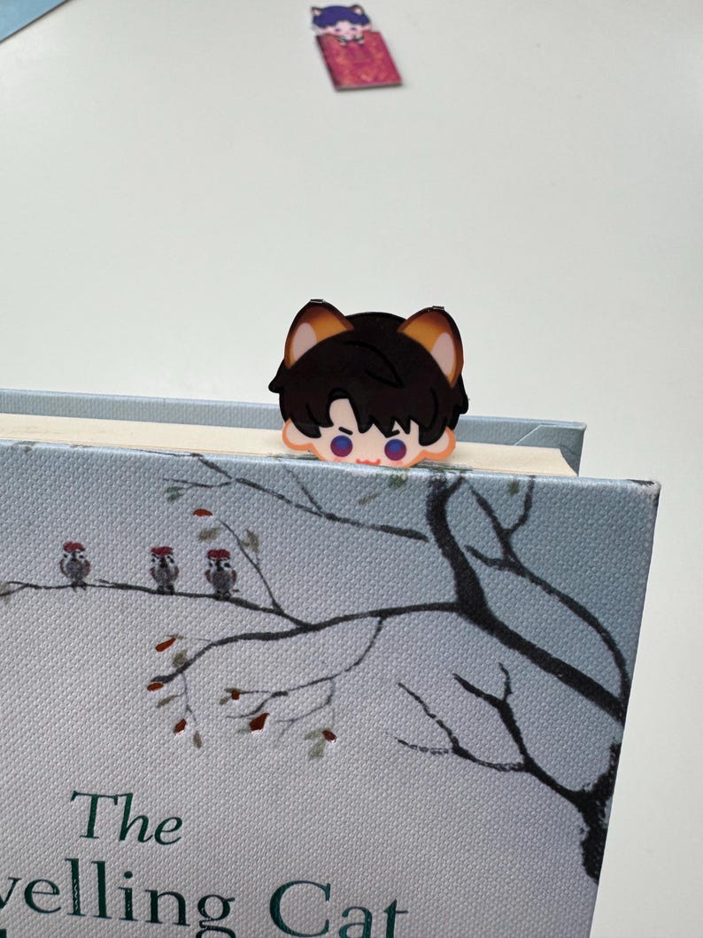 May include: A black and brown anime-style bookmark in the shape of a character's head, placed on top of a book. The book has a light blue cover with a tree branch design and the title "The Travelling Cat".