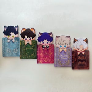 May include: A collection of five decorative bookmarks featuring anime-style characters with cat ears. Each character is perched atop a miniature book with titles like "Frozen," "Peter Pan," and "Dracula." The bookmarks have a cute and whimsical design.