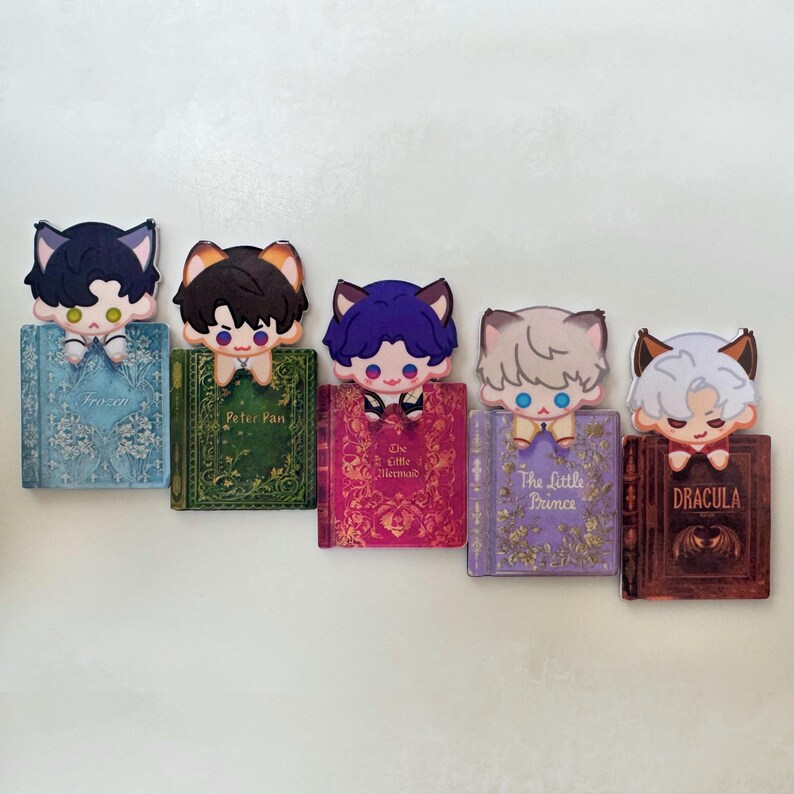 May include: A collection of five decorative bookmarks featuring anime-style characters with cat ears. Each character is perched atop a miniature book with titles like "Frozen," "Peter Pan," and "Dracula." The bookmarks have a cute and whimsical design.