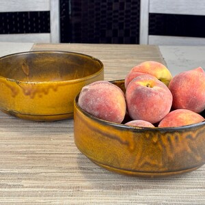 May include: Two round ceramic bowls with a deep mustard yellow and dark brown drip glaze finish. One bowl is empty, while the other is filled with several fresh, fuzzy peaches with pink and orange skin tones, showcasing a smooth, rounded shape.