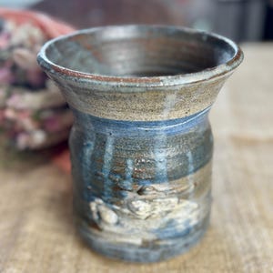 May include: Handmade ceramic vase with a wide, flared rim. The vase features a textured surface with shades of blue, brown, and beige. The design includes a band of blue near the middle and a rough, organic texture.