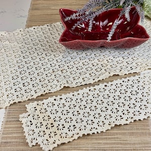 May include: Two white crocheted table runners with a delicate floral pattern. The runners are on a brown woven mat.