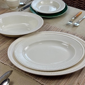 May include: A collection of off-white ceramic dinnerware, including oval platters, bowls, and plates. The pieces have a ribbed design around the edges and are arranged on a neutral-toned tablecloth. Silverware and a teacup are also visible.