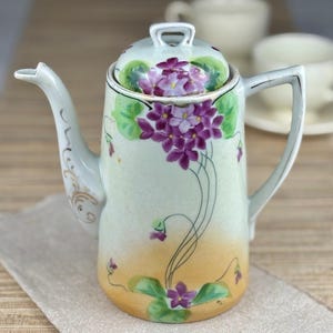 May include: A vintage porcelain teapot with a floral design. The teapot is decorated with purple flowers and green leaves on a light green background. The lid has a gold trim.