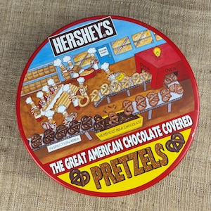May include: A round tin featuring a cartoon illustration of a pretzel factory. The tin is red with yellow and white text that reads "HERSHEY'S" and "THE GREAT AMERICAN CHOCOLATE COVERED PRETZELS." The illustration shows bakers and pretzels.