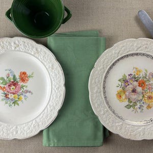 May include: Two white plates with floral patterns and scalloped edges, a green bowl, and a green napkin are arranged on a neutral surface. A knife and fork are placed on the right. The plates feature colourful floral designs.