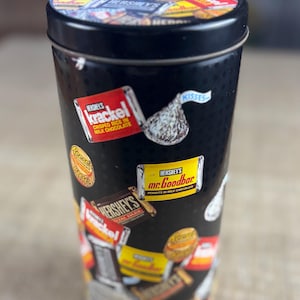 May include: A black cylindrical tin with a lid, covered in colorful Hershey's candy labels. The labels include "Krackel," "Mr. Goodbar," and "Kisses." The lid is also covered in candy labels. The tin is likely used for storage or as a collectible item.