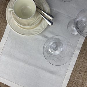 May include: Cream-colored teacup and saucer set with a matching bowl and two silver spoons. The tableware is arranged on a white linen runner, alongside clear etched glassware. The runner is placed on a burlap surface.