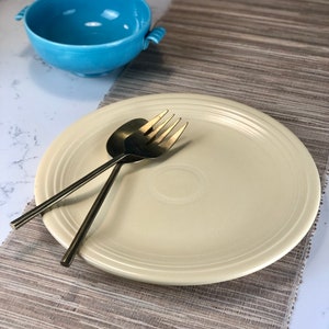 May include: A pale yellow ceramic dinner plate with a raised rim and a subtle pattern. Two gold-toned forks are placed on the plate. The plate is on a brown woven placemat.