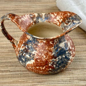 May include: A ceramic jug with a wide spout and a curved handle. The jug features a speckled pattern in shades of brown, blue, and white. The interior is a light beige colour. This decorative jug is suitable for serving beverages.