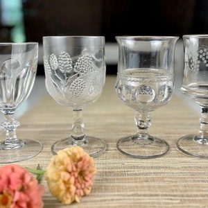 May include: Four clear glass goblets with etched designs. The goblets have different patterns, including grapes, berries, and geometric shapes. The goblets are arranged on a woven placemat.