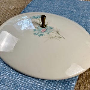May include: An oval-shaped, off-white ceramic lid featuring a central floral pattern with light blue petals and a small, dark metallic knob handle. The surface is smooth and glossy, reflecting light across its curved, rounded top edge.
