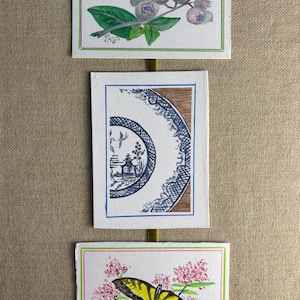 May include: Three watercolor paintings on white paper, each with a green and pink border. The top painting depicts blueberries and leaves. The middle painting shows a blue and white plate design. The bottom painting features a yellow butterfly.