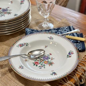 May include: A stack of white porcelain soup bowls with floral designs, a silver spoon, and a clear glass. The bowls have a gold rim and a floral pattern with pink, purple, and green flowers. A blue patterned napkin and a knife are also visible.