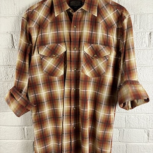 Peut inclure: Une chemise à carreaux à manches longues en brun, beige et blanc avec un col boutonné et deux poches poitrine. La chemise porte l'étiquette de la marque Pendleton sur le côté gauche de la poitrine.