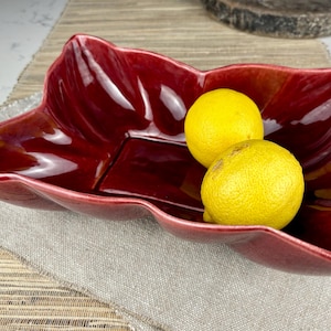 May include: A red ceramic bowl shaped like a leaf with two yellow lemons inside.