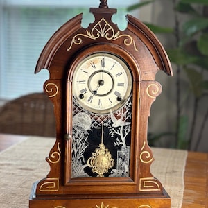 May include: A wooden mantel clock with a dark brown finish and gold painted scrollwork accents. The clock face features Roman numerals and black hands. Below the face, a glass panel displays a white etched nature scene with a bird and a gold pendulum.