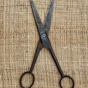 May include: Antique metal scissors with pointed blades and circular finger holes. The scissors have a dark, aged appearance, suggesting they are vintage or well-used. The blades are open, displaying their sharp edges. The scissors are set against a textured, beige background.