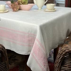 May include: A white linen tablecloth with pink stripes and a delicate floral pattern. The tablecloth is laid on a table with a white teapot, two teacups, and a pink bowl filled with flowers.