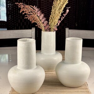 May include: Three off-white ceramic vases with a bulbous base and cylindrical neck. One vase holds dried flowers and grasses. The vases are arranged on a beige woven placemat. The background includes a dark brown screen and two light-colored chairs.