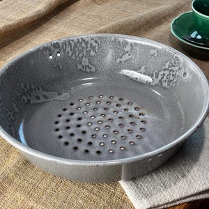 May include: A gray ceramic colander with a series of small holes in the bottom. The colander has a wide rim and a slightly textured surface. A green teacup and saucer are in the background.