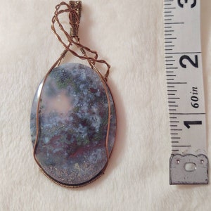 May include: A copper wire wrapped pendant featuring a light gray, pink, and green agate stone. The stone is oval shaped and measures approximately 1.5 inches in length.