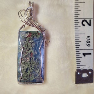 May include: A silver wire wrapped pendant featuring a rectangular moss agate stone with green, blue, and red patterns. The pendant measures approximately 1 inch in length.