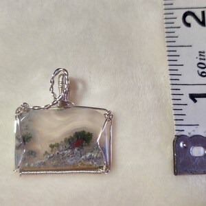 May include: A silver wire wrapped pendant featuring a clear agate stone with green and red inclusions. The pendant is approximately 1 inch in length.