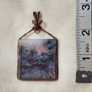 May include: A square-shaped agate pendant with a copper wire frame. The agate has a variety of colors, including pink, white, gray, and green. The pendant is approximately 1.5 inches in size.