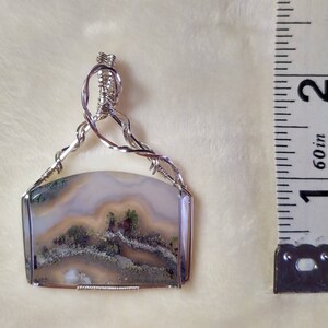 May include: A silver wire wrapped pendant featuring a light brown, white, and green agate stone with a landscape pattern. The pendant is approximately 1 inch in length.