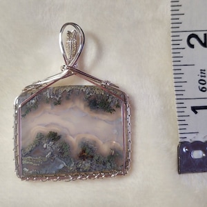 May include: A silver wire wrapped pendant featuring a square, moss agate stone with a white, gray, and green pattern. The pendant is approximately 1.5 inches in size.