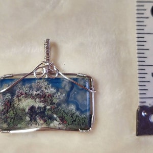 May include: A silver wire wrapped pendant featuring a blue, green, and white agate stone with a mossy pattern. The pendant is approximately 1 inch in length.