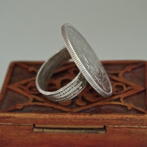 Può includere: Un anello d'argento con un design a moneta. L'anello è su una scatola di legno con intagli intricati.