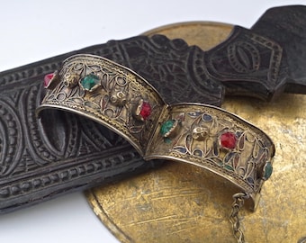 May include: A silver bracelet with intricate filigree work and red and green gemstones. The bracelet is on a dark brown surface.