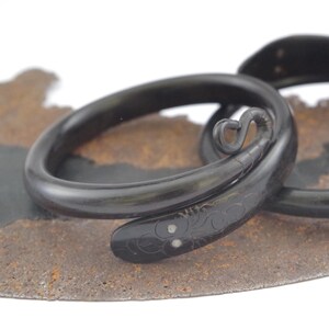 Può includere: Un bracciale nero a spirale a forma di serpente con una finitura liscia e lucida. La testa del serpente presenta motivi a scaglie incisi e due piccoli inserti circolari di colore chiaro per gli occhi, mentre la coda termina con un piccolo ricciolo.