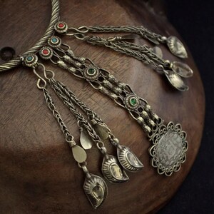 Op de afbeelding: Een antieke zilverkleurige ketting met ingewikkelde details. De ketting heeft een gedraaide kraag, decoratieve kettingen en bungelende hangers. Kleine, kleurrijke edelstenen in rood, groen en blauw zijn in het ontwerp gezet. De ketting wordt op een houten oppervlak gepresenteerd.