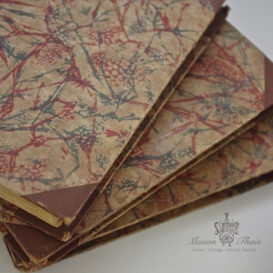 Könnte beinhalten: Ein Stapel antiker Bücher mit braunen Lederrücken und gemusterten Papierumschlägen. Die Umschläge zeigen ein rotes und blaues Blumenmuster auf beigefarbenem Hintergrund.