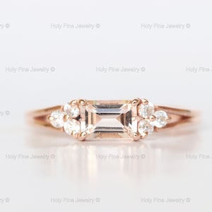May include: A rose gold engagement ring with a rectangular center stone and smaller round stones on either side.