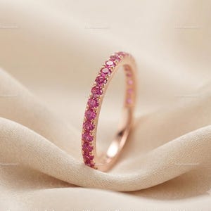 May include: A thin rose gold band ring featuring a continuous row of small, round, vibrant pink gemstones set along the outer surface. The metal has a polished finish, and the stones are arranged in a classic eternity style setting.