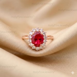 Könnte beinhalten: Ein Ring aus Roségold mit einem großen, oval geschliffenen roten Mittelstein, der von einem Kranz aus kleinen, klaren, rund geschliffenen Steinen umgeben ist. Die Ringschiene ist schmal und poliert und zeigt ein klassisches Design.