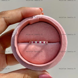 Può includere: Un delicato anello con una fila di piccole pietre preziose rosse e diamanti bianchi incastonati in una fascia in oro rosa. L'anello è presentato in una scatola per anelli in velluto rosa.