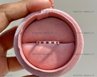Könnte beinhalten: Ein zarter Ring mit einer Reihe kleiner roter Edelsteine und weißen Diamanten, die in einer Roségold-Schiene gefasst sind. Der Ring wird in einer rosafarbenen Samt-Ringbox präsentiert.