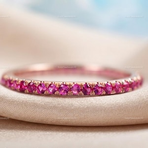Könnte beinhalten: Ein Ring aus Roségold mit einer durchgehenden Reihe kleiner, runder, leuchtend pinker Edelsteine, die entlang der Außenfläche gefasst sind. Das Metall hat ein warmes, metallisches Finish und die Steine sind einheitlich groß, was ein funkelndes Design ergibt.