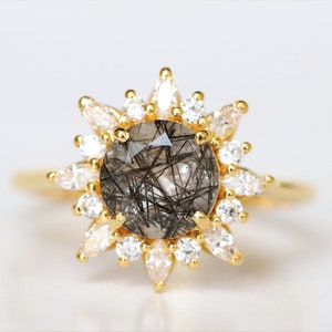 Könnte beinhalten: Ein goldener Ring mit einem großen, runden, schwarzen und klaren Edelstein, der von kleineren, klaren Edelsteinen umgeben ist.
