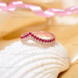 May include: Rose gold chevron ring with alternating red and clear gemstones. The ring is set on a white seashell, with a pink ribbon in the background. The ring is a piece of fine jewellery.