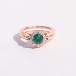 Könnte beinhalten: Ein Ring aus Roségold mit einem grünen Edelstein, der von einem Heiligenschein aus kleineren, klaren Edelsteinen umgeben ist. Der Ring ist mit dem Text "Holyfire Jewelry" graviert.
