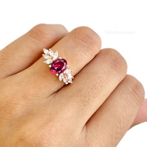 May include: A gold ring with a large oval-shaped red gemstone surrounded by smaller white gemstones. The ring is set on a finger.